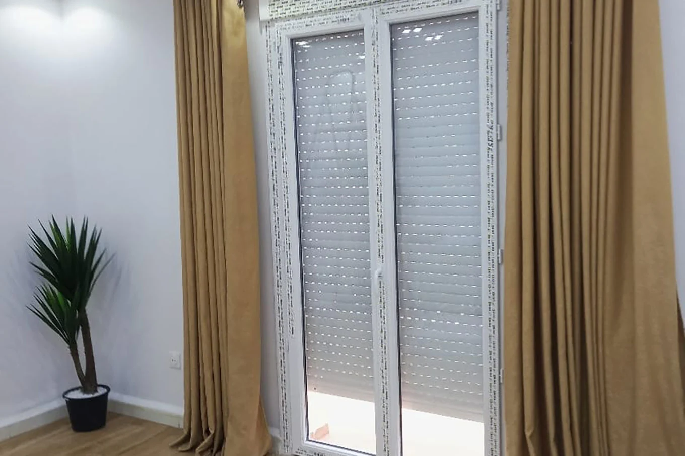 porte fenetre pvc blanche avec un volet roulant en lame pvc presque fermé amenagé d'un cache rideau marron dans une salon décoré avec une plante